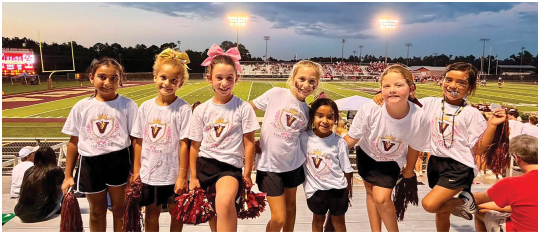 Seven children wearing matching cheerleading outfits with pom-poms stand together smiling on bleachers at a sports field under a cloudy evening sky. Description generated by AI