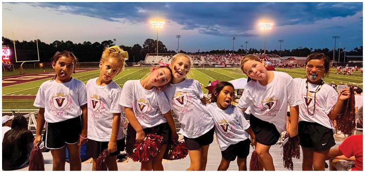A group of children wearing white shirts and colorful bows are standing close together on a sports field at dusk with stadium lights and bleachers in the background. Description generated by AI