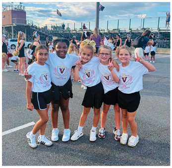 Five children wearing matching cheerleading shirts and black skirts or shorts stand together outdoors with their arms around each other, smiling and making peace signs. Description generated by AI
