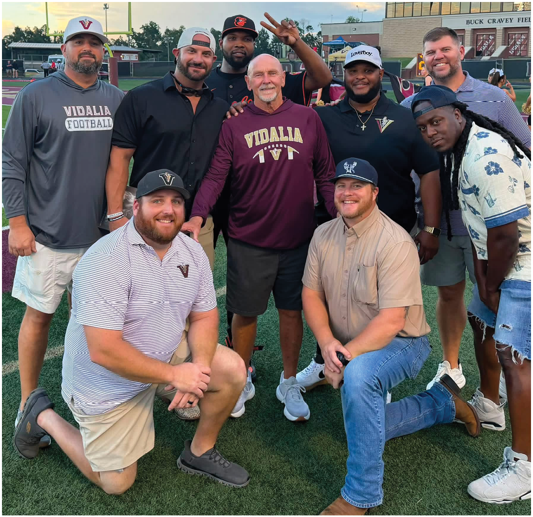 Two people wearing casual clothing and baseball caps are kneeling on a sports field while several others stand behind them. Description generated by AI