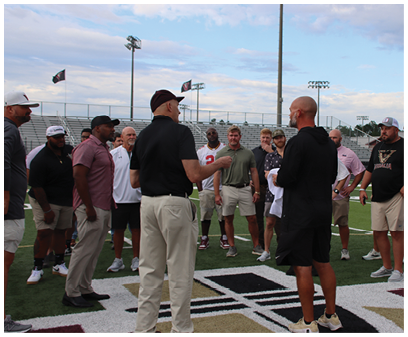 A group of adults standing and talking on a sports field near bleachers under a partly cloudy sky. Description generated by AI