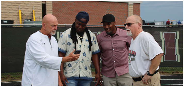Four adults stand outdoors near a fence with one person showing something on a phone to the others, who are attentively looking at the screen. Description generated by AI