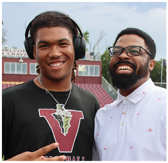 Two adults are smiling and posing together outdoors, one wearing headphones and a black shirt with a large “V" and the other wearing glasses and a white button-up shirt with small red patterns. Description generated by AI
