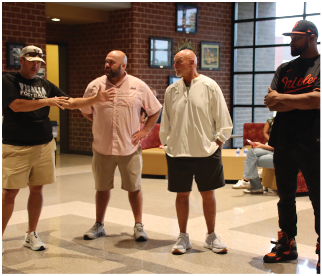 Four adults stand indoors on a polished floor with brick walls and windows in the background, wearing casual clothing including shorts, sneakers, and a sports jersey. Description generated by AI