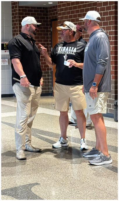 Three adults wearing casual clothing and sneakers are standing and conversing outside near a brick wall and a trash bin. Description generated by AI