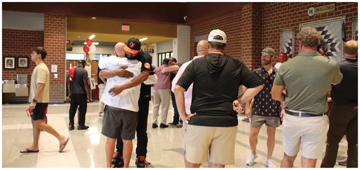 People in casual clothing are hugging and standing in a brightly lit indoor space with brick walls and tiled floors. Description generated by AI