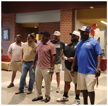 A group of seven adults wearing casual clothing and hats stand indoors in a semi-circle, some holding drinks, with a brick wall and framed artwork in the background. Description generated by AI