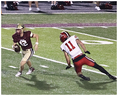 Two people in football uniforms are on a field with one holding a football and the other approaching in a defensive stance. Description generated by AI