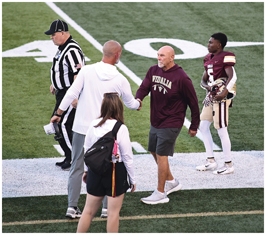 Two adults shake hands on a football field near a referee, a player in uniform holding a helmet, and a person with a backpack walking nearby. Description generated by AI