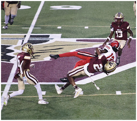 Four people in football uniforms are on a field with one player in red being tackled mid-air by a player in maroon and gold. Description generated by AI