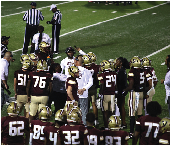 A group of football players in maroon and gold uniforms gather on the field with coaches and referees during a game. Description generated by AI