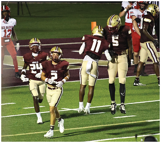 Several people wearing football uniforms are standing and walking on a green field under stadium lights. Description generated by AI