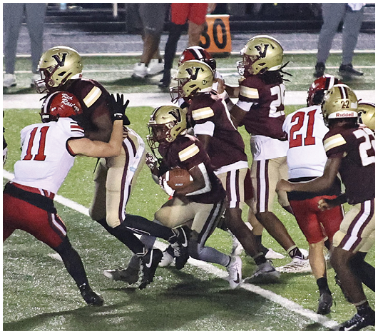 People wearing football uniforms are engaged in a play on a grassy field with one person holding the ball while others attempt to block or tackle. Description generated by AI