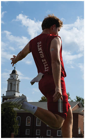 A person wearing a red athletic outfit with “LITTLE GIANTS" on the back is captured mid-jump outdoors near a building with a clock tower against a partly cloudy sky. Description generated by AI