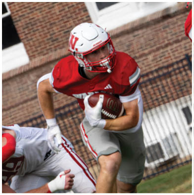 A group of people wearing red and white football uniforms are actively engaged in a game on a grassy field with one person holding a football tightly while others attempt to block or tackle. Description generated by AI