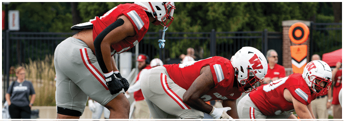 A person wearing a red football uniform with the number seven stands on a field with a crowd of people in the background and another person in a white football uniform nearby. Description generated by AI