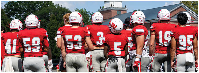 Players in red and white football uniforms are lined up on the field facing each other, preparing for a play during a game with spectators in the background. Description generated by AI