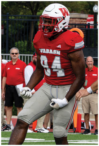 Two adults in football uniforms are jumping and reaching for the ball during a game with other players and a person in casual clothing in the background. Description generated by AI