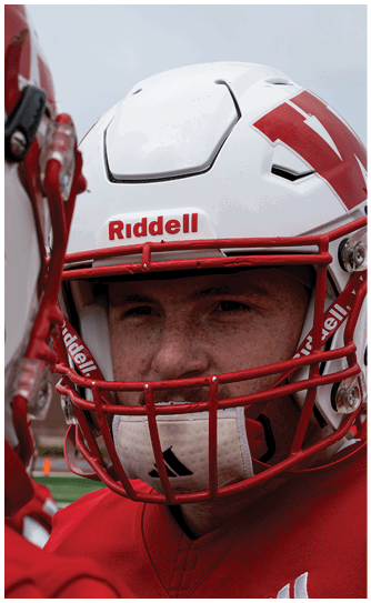 A person in a red Wabash football uniform is running on a field while holding a football, with other players and spectators visible in the background. Description generated by AI
