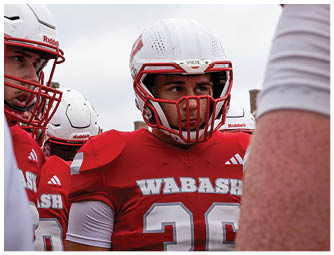 A group of people in red and white football uniforms are lined up on a green field preparing for a play with a large brick building and spectators in the background. Description generated by AI