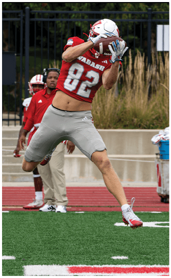 A person in a red Wabash football uniform is running on a field while holding a football, with other players and spectators visible in the background. Description generated by AI