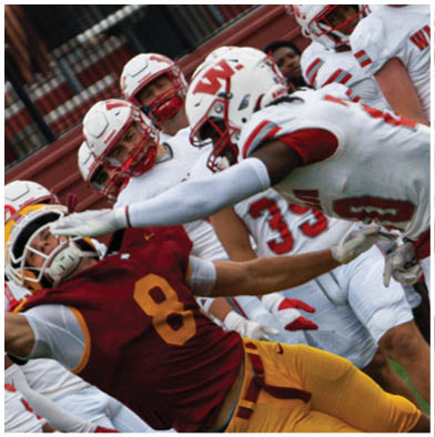 A group of people wearing red and white football uniforms are actively engaged in a game on a grassy field with one person holding a football tightly while others attempt to block or tackle. Description generated by AI