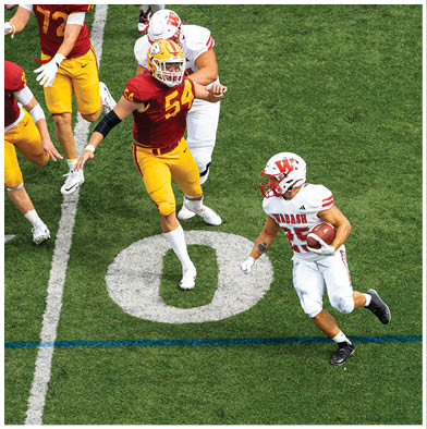 People wearing red and white striped overalls are gathered outdoors with one holding a red flag that says Wabash while a crowd watches from bleachers in the background. Description generated by AI