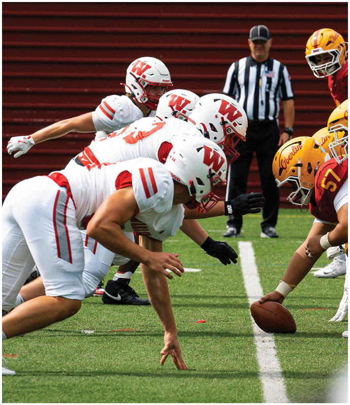 A person in a red football uniform tackles another person in a white football uniform who is holding a football on a green field during a game. Description generated by AI