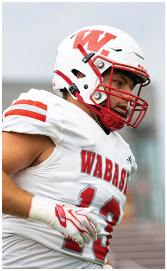 A person in a red Wabash football uniform is running on a field while holding a football, with other players and spectators visible in the background. Description generated by AI