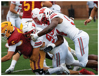 A group of people in red and white football uniforms are lined up on a green field preparing for a play with a large brick building and spectators in the background. Description generated by AI