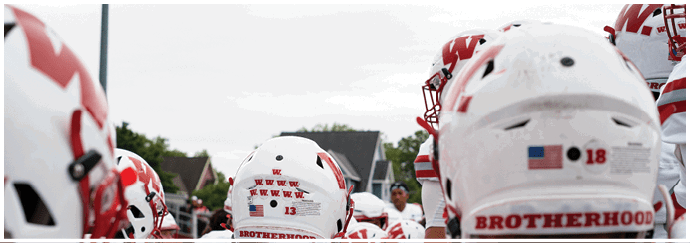 A person wearing a red football uniform with the number seven stands on a field with a crowd of people in the background and another person in a white football uniform nearby. Description generated by AI