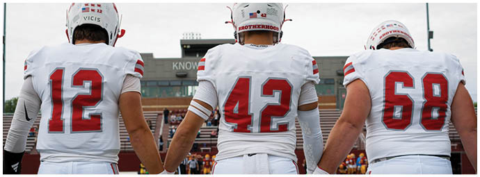 Players in red and white football uniforms are lined up on the field facing each other, preparing for a play during a game with spectators in the background. Description generated by AI