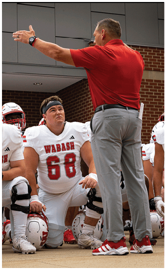 A person in a red Wabash football uniform is running on a field while holding a football, with other players and spectators visible in the background. Description generated by AI