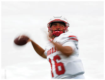 A group of people in red and white football uniforms are lined up on a green field preparing for a play with a large brick building and spectators in the background. Description generated by AI