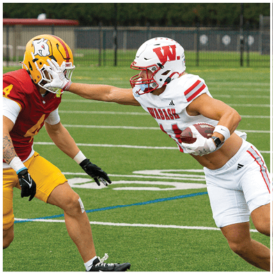 Four people in football uniforms are on a field with one person holding a football while others are attempting to tackle or block. Description generated by AI