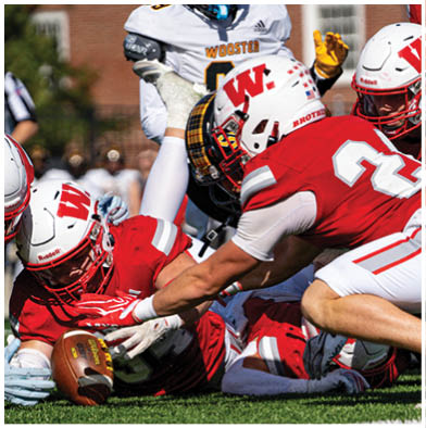A group of people wearing red and white football uniforms are actively engaged in a game on a grassy field with one person holding a football tightly while others attempt to block or tackle. Description generated by AI