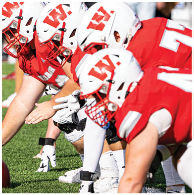 People wearing red and white striped overalls are gathered outdoors with one holding a red flag that says Wabash while a crowd watches from bleachers in the background. Description generated by AI