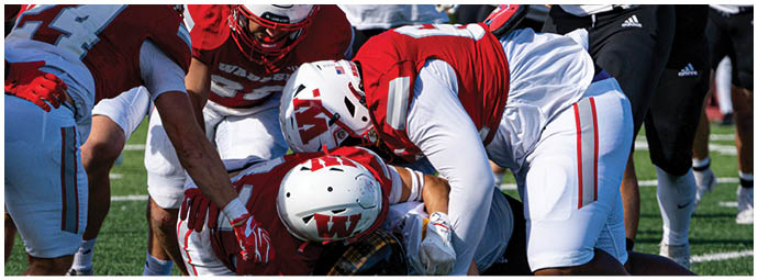 A group of people wearing white helmets with red “W" logos and red sports uniforms are huddled together on a green field. Description generated by AI