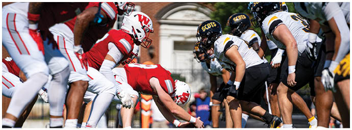 Players in red and white football uniforms are lined up on the field facing each other, preparing for a play during a game with spectators in the background. Description generated by AI