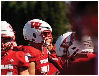 A group of people in red and white football uniforms are lined up on a green field preparing for a play with a large brick building and spectators in the background. Description generated by AI