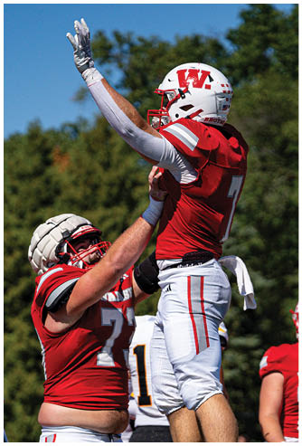 Two adults in football uniforms are jumping and reaching for the ball during a game with other players and a person in casual clothing in the background. Description generated by AI