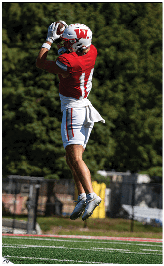 A person in a red Wabash football uniform is running on a field while holding a football, with other players and spectators visible in the background. Description generated by AI