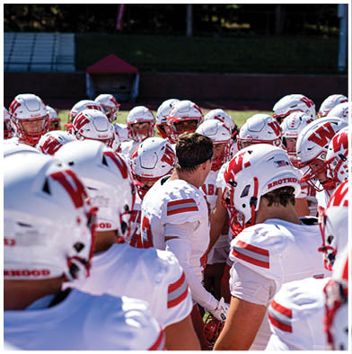 Four people in football uniforms are on a field with one person holding a football while others are attempting to tackle or block. Description generated by AI