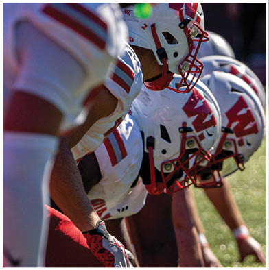People wearing red and white striped overalls are gathered outdoors with one holding a red flag that says Wabash while a crowd watches from bleachers in the background. Description generated by AI