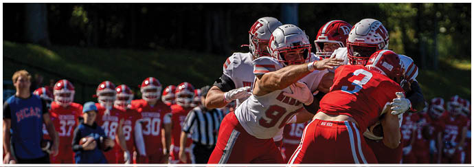 A person wearing a red football uniform with the number seven stands on a field with a crowd of people in the background and another person in a white football uniform nearby. Description generated by AI