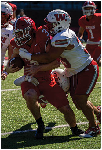Two adults in football uniforms are jumping and reaching for the ball during a game with other players and a person in casual clothing in the background. Description generated by AI