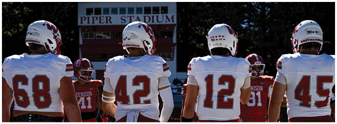 Players in red and white football uniforms are lined up on the field facing each other, preparing for a play during a game with spectators in the background. Description generated by AI
