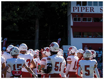 A group of people in red and white football uniforms are lined up on a green field preparing for a play with a large brick building and spectators in the background. Description generated by AI
