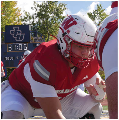 People wearing red and white striped overalls are gathered outdoors with one holding a red flag that says Wabash while a crowd watches from bleachers in the background. Description generated by AI