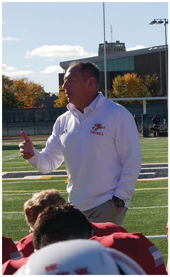 A person in a red Wabash football uniform is running on a field while holding a football, with other players and spectators visible in the background. Description generated by AI